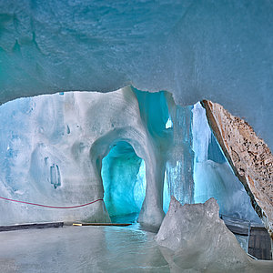 Eine Höhle aus blauem Eis mit glatten, abgerundeten Wänden, einer tunnelartigen Öffnung und einem Holzsteg auf dem Eisboden.