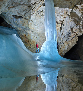 Ein Höhlenführer in einer roten Jacke steht in der Höhle und blickt zu einer großen gefrorenen Eisformation hinauf, die von Eis und Felswänden umgeben ist, auf deren eisigem Boden sich Spiegelungen zeigen.