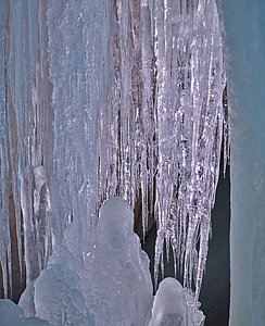 Lange, klare Eiszapfen hängen dicht aneinander, an deren Basis sich Hügel aus gefrorenem Eis bilden. Die Szene hat einen kalten, bläulichen Farbton und erzeugt eine winterliche und eisige Atmosphäre.