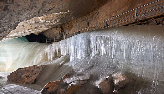 Eine große Eisschicht bedeckt den Höhlenboden. Ein Metallgeländer verläuft entlang der Felswand über dem Eis, und die Höhlendecke erhebt sich darüber.