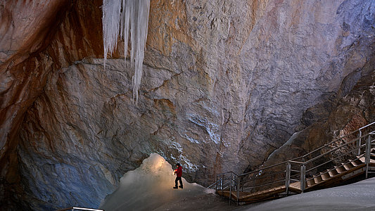 Eine Person in einer roten Jacke steht neben einer Treppe in einer großen Höhle mit Eiswänden und riesigen Eiszapfen, die von der Decke hängen.