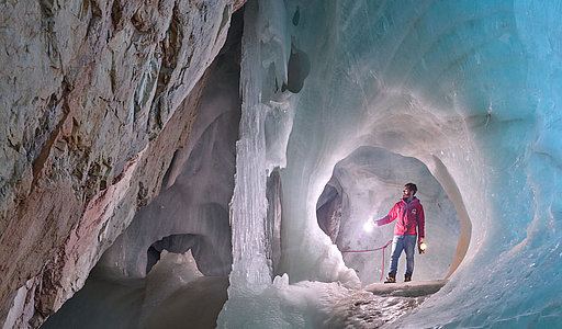 Ein Höhlenführer in roter Jacke steht mit einer Lampe in der Hand in der Eishöhle. Dicke Eisformationen und glatte, bläulich gefärbte Wände umgeben ihn.
